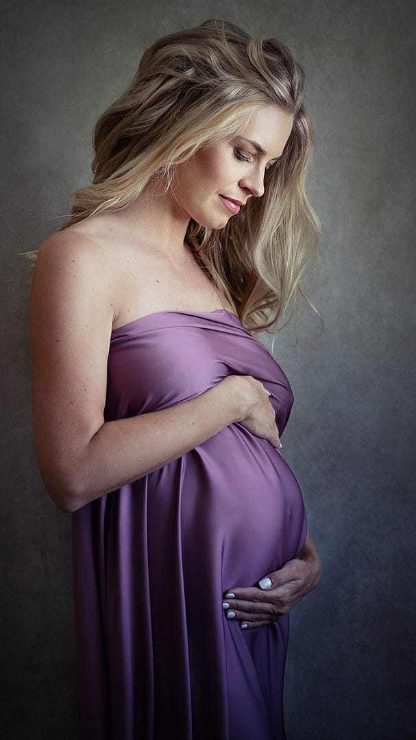 Certainly: **Alt**: Maternity photo in Dubai of a blond pregnant woman draped in shiny purple cloth, gazing at her belly.