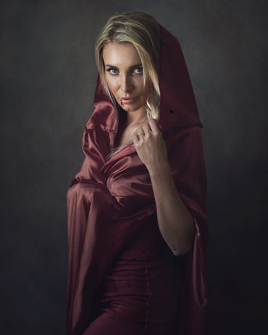 Artistic portrait photography brings to life a fine art portrait of a blonde woman, gracefully draped in a red cape with a subtle blood drip from her lips, embodying a painterly style and encapsulating a mysterious Halloween aura.